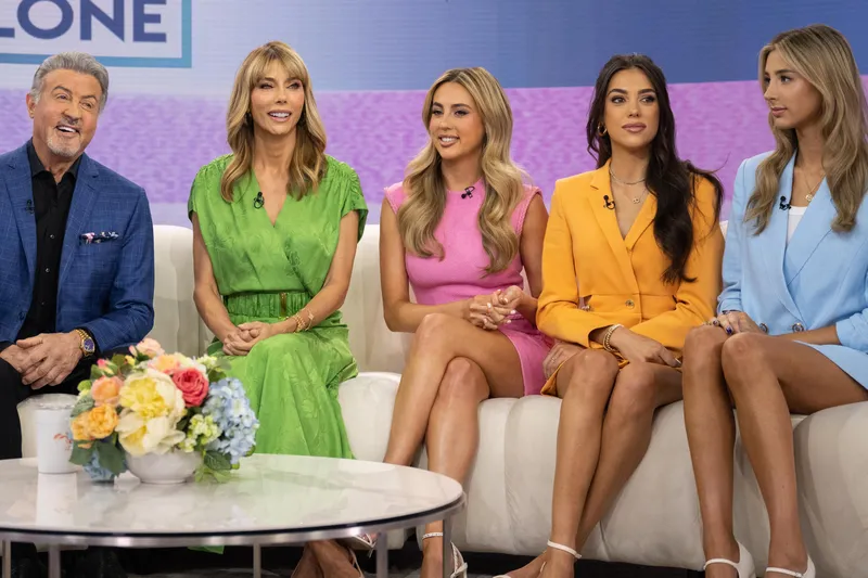 Sylvester Stallone and Jennifer Flavin on white sofa, three daughters Sistine, Sophia, and Scarlet seated beside them to right; round table at center; blue, purple, fuchsia striped background