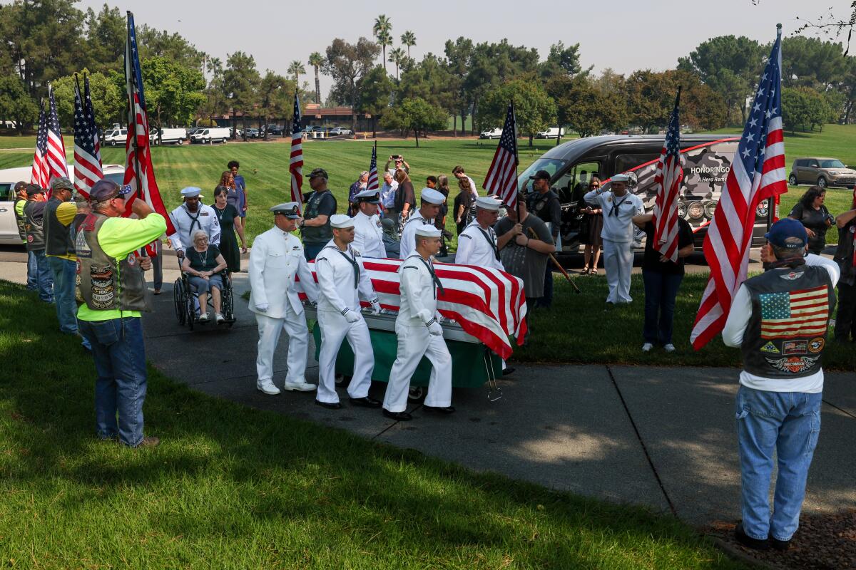 A photo of sailors carrying a casket in a cemetery as people hold American flags and salute.