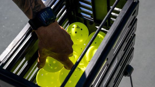 A hand reaches into a bin of pickleballs