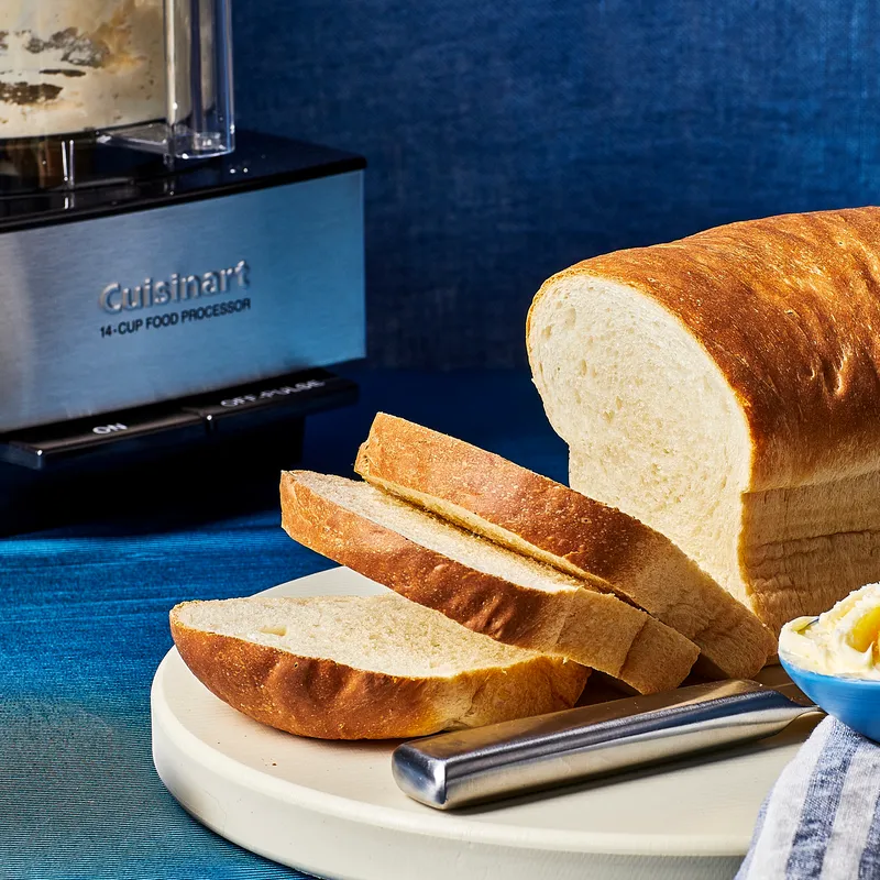 Sliced white bread on stone surface in a blue fabric backdrop with a cuisine art food processor in the back