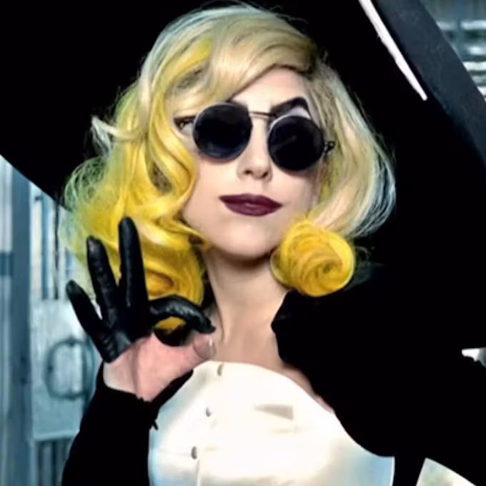 A woman with glamorous wavy hair featuring yellow highlights wears dark sunglasses and black gloves, striking a playful pose against a modern backdrop.