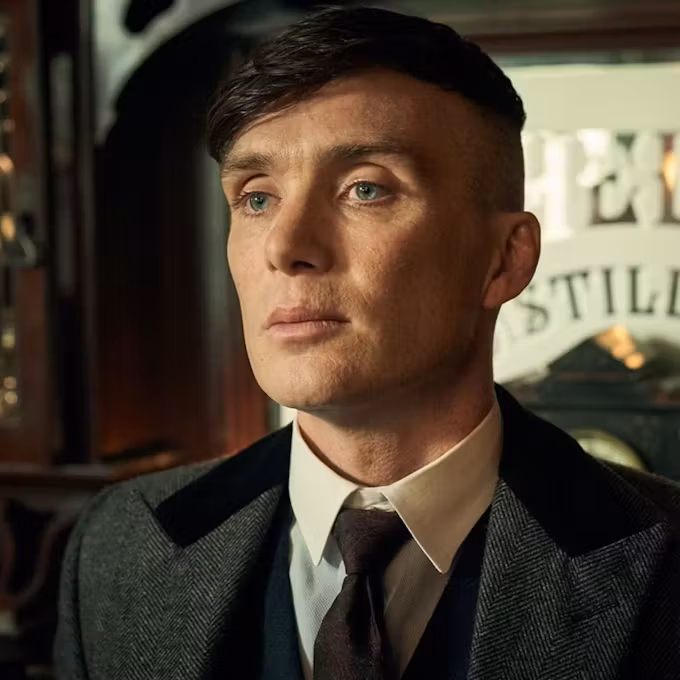 A well-dressed man with a serious expression stands in front of a vintage distillery backdrop, exuding an air of confidence and intensity.