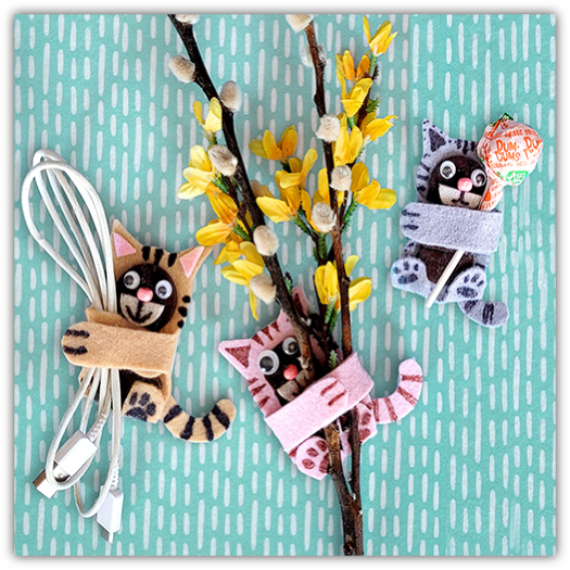Crafty felt cat huggers on a flowering branch, featuring three cute cat designs: one in tan with a whisk, one in gray wrapped in a scarf, and one in pink, all decorated with playful patterns and placed against a teal background with a raindrop pattern.