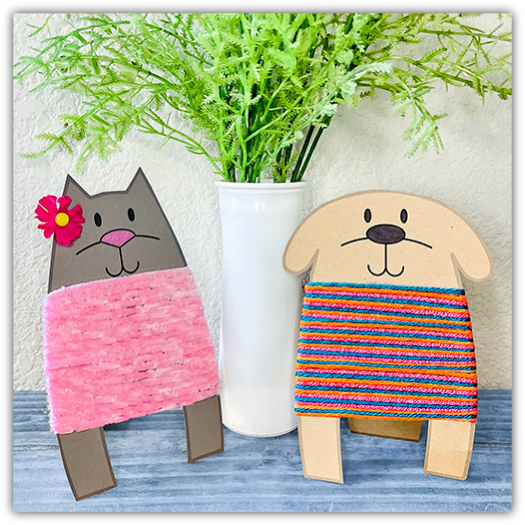 Two adorable craft figures made of cardboard and yarn, depicting a cat and a dog, positioned on a table next to a white vase with green plants. The cat figure is wrapped in pink yarn with a flower on its ear, while the dog figure is covered in colorful striped yarn, both showcasing cheerful and playful designs.