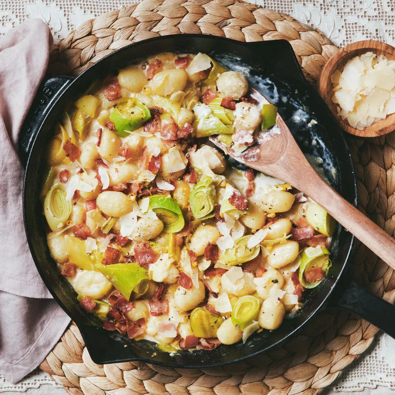 A cast-iron skillet with sautéed shelf-stable gnocchi, crispy bacon, leeks, and parmigiana in a creamy sauce and topped with cracked black pepper.
