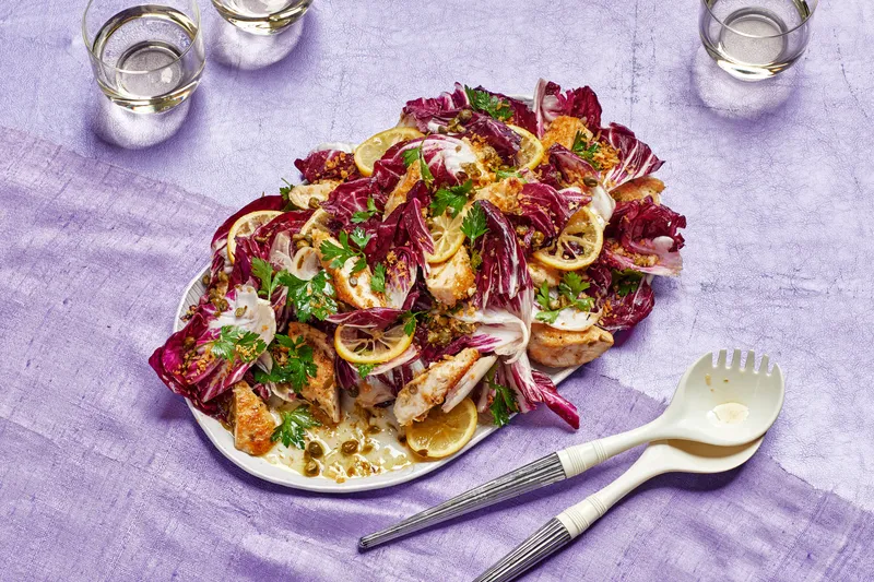Platter of warm chicken piccata salad with radicchio, parsley, and capers, topped with sliced lemons