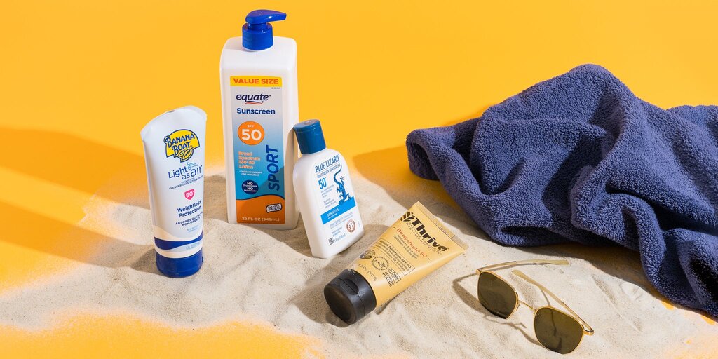 Four different sunscreens sit on a pile of sand next to a dark blue towel. The whole scene is set upon a yellow background.