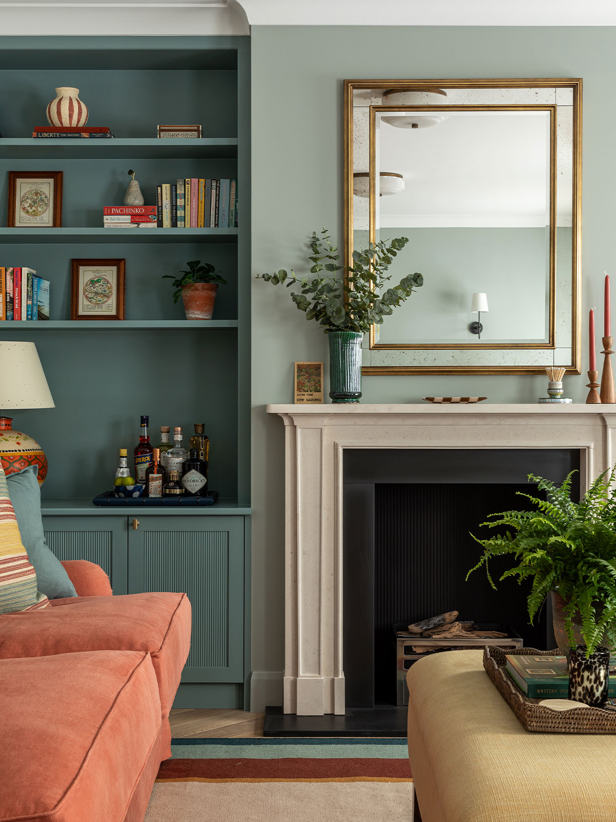 A classic living room with a fireplace on one wall, a large, gold-framed mirror above the mantle and a built-in storage unit to one side. The room is painted in shades of blue, creating a calm, breezy feel, like looking out onto a peaceful sea. There’s also a squishy orange sofa in the room, adding a vibrant contrast and playful element.