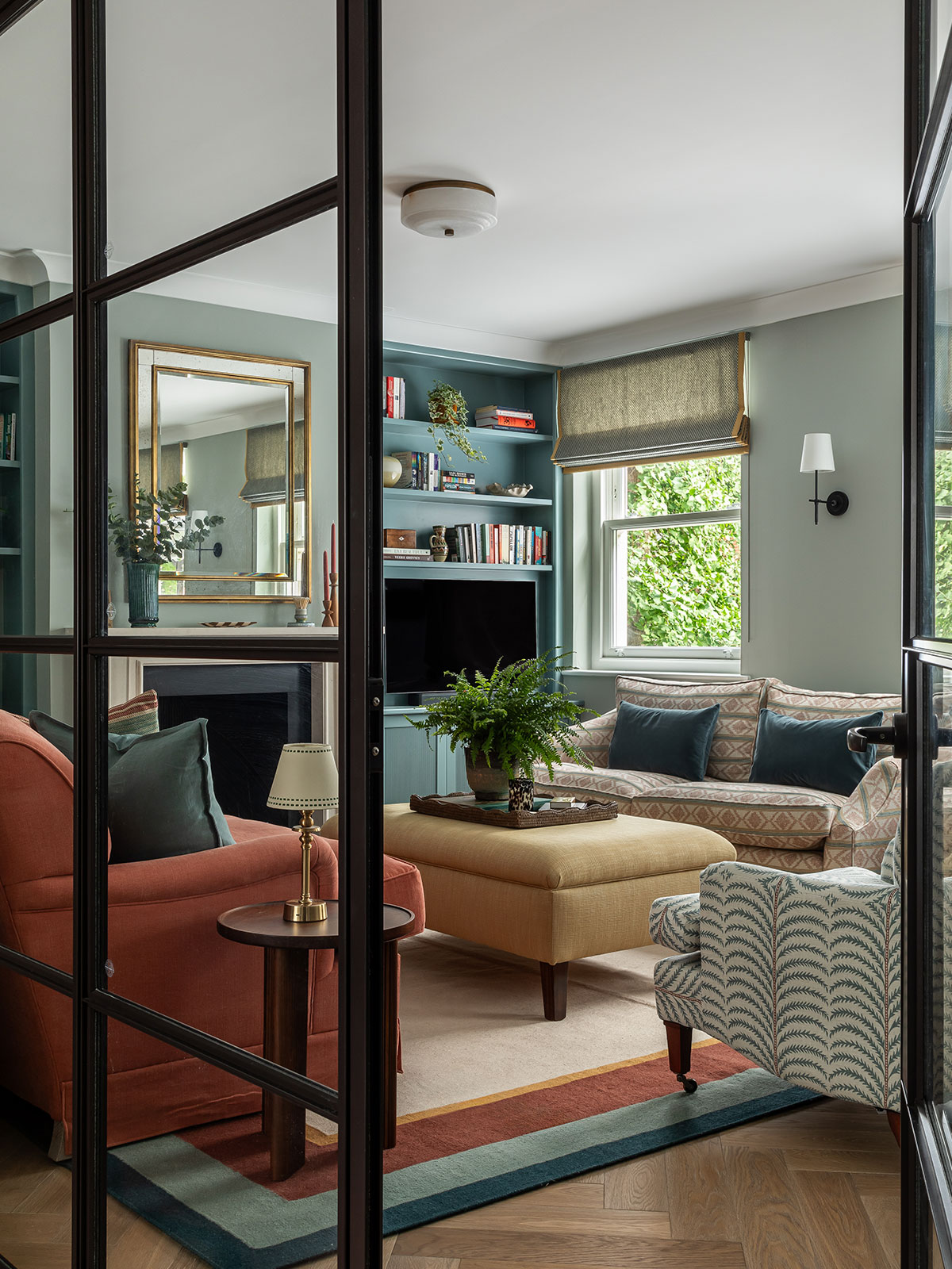 The view through an open, Crittall glass door into a playful but sophisticated living room. There’s a fireplace on one wall with a large, gold-framed mirror above the mantle and built-in storage units on either side containing books, a TV and ornaments. The room is painted in shades of blue, creating a calm, breezy feel, like looking out onto a peaceful sea. There’s also two squishy, upholstered sofas and a chair in a mixture of colours and patterns, creating a playful, eclectic look.