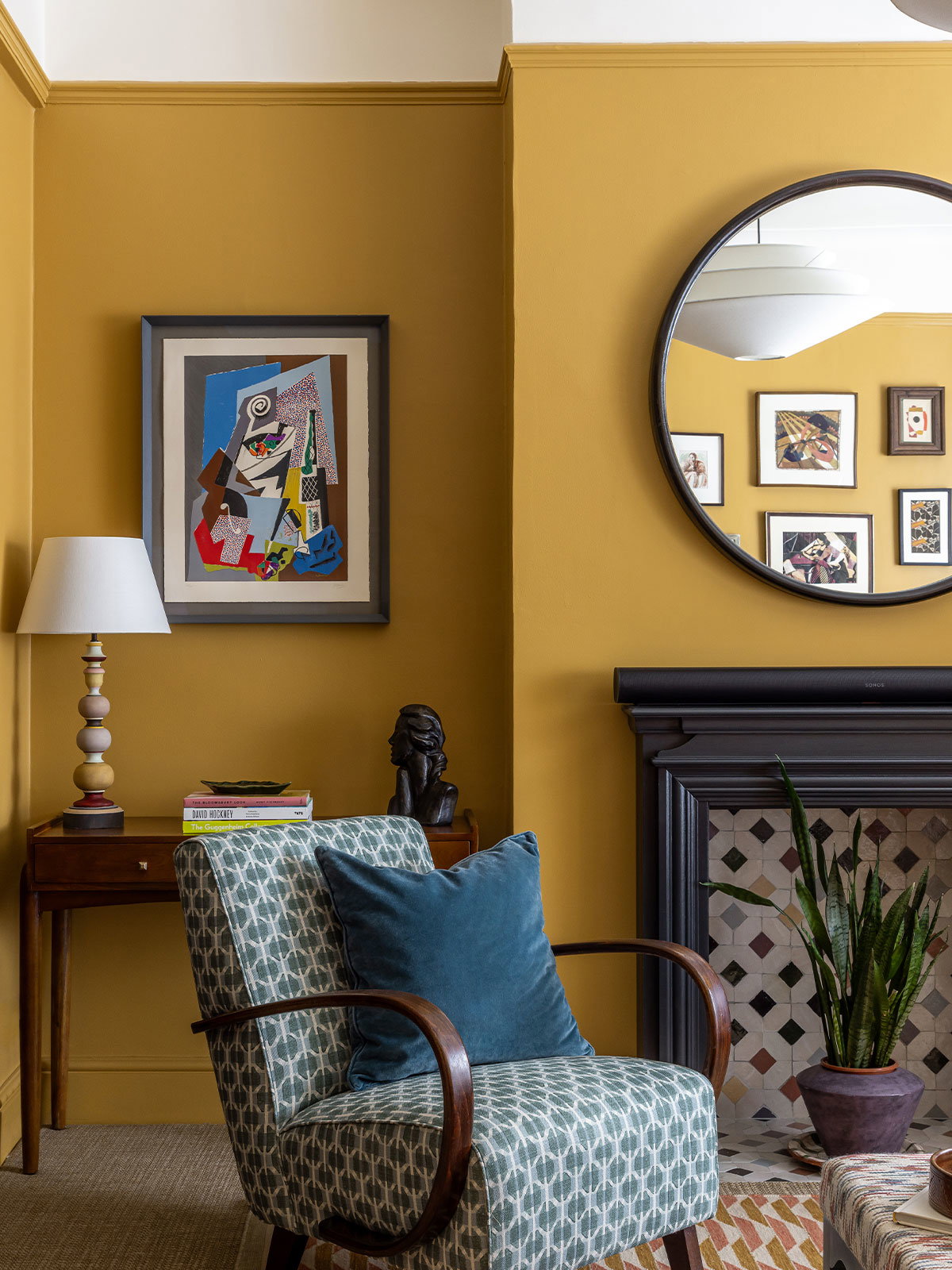 A warm, inviting living room with a traditional fireplace and a large, round mirror above it. The bottom two thirds of the walls are painted in a rich yellow shade with a crisp white on the top third and ceiling. The contrast between the two shades makes the room feel bright and airy, while also cosy and comfortable. In front of the fireplace is a mid-century modern style chair.