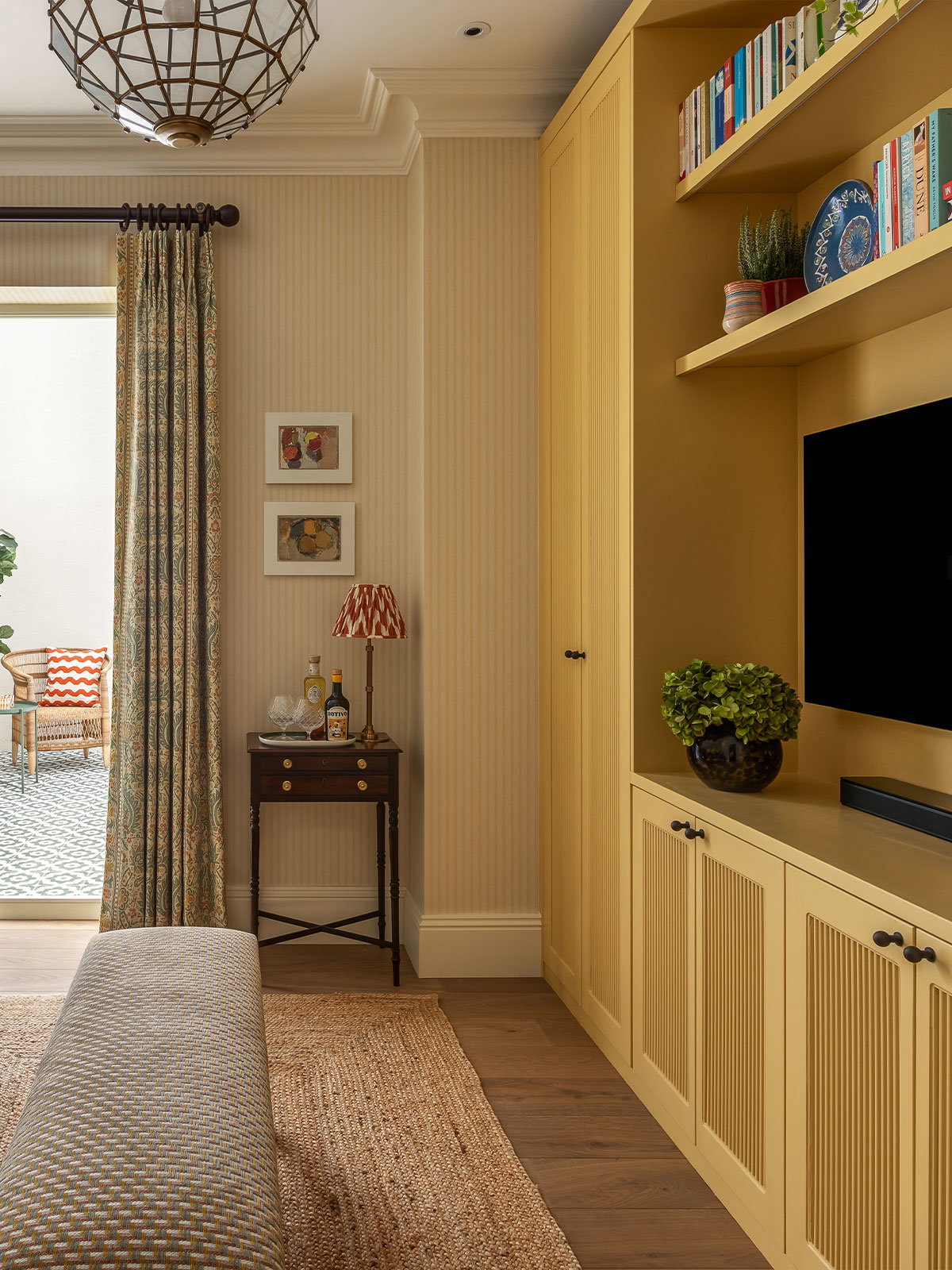 A light, airy living room with doors opening to a bright garden room. The living room has a traditional, country feel with striped wallpaper, jute rug and plump blue sofa with comfortable, plump pillows. The space invites you to sit down and relax.