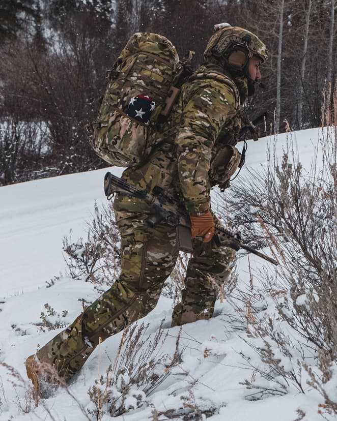 Operator hiking a snowy hillside while carrying a rifle.