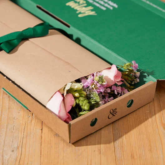 Pink, purple and cream flowers can be seen poking out the top of carefully packaged floral wrap, tied with a green ribbon in Bloom & Wild letterbox packaging.