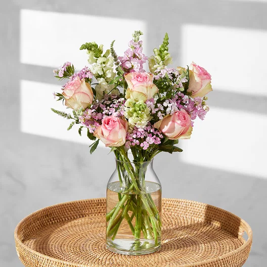 A gorgeous bouquet of pink, cream and purple made up of snapdragons, roses, stocks and dianthus sits in a clear vase on a wicker tray.
add the text here.