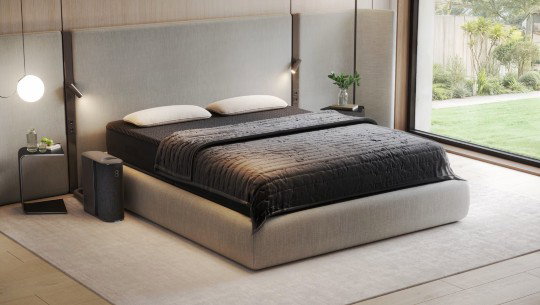 A bed for two with dark sheets and a bedside console resembling a large router