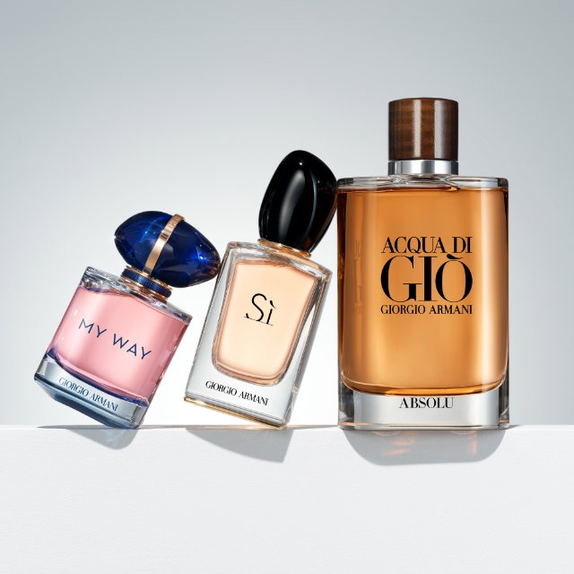 Three Giorgio Armani perfume bottles are arranged diagonally on a white surface against a light grey background: "My Way" (pink liquid, blue organic cap) leaning against "Sì" (amber liquid, black organic cap), which in turn leans against Acqua di Giò Absolu amber liquid, wooden-textured cap