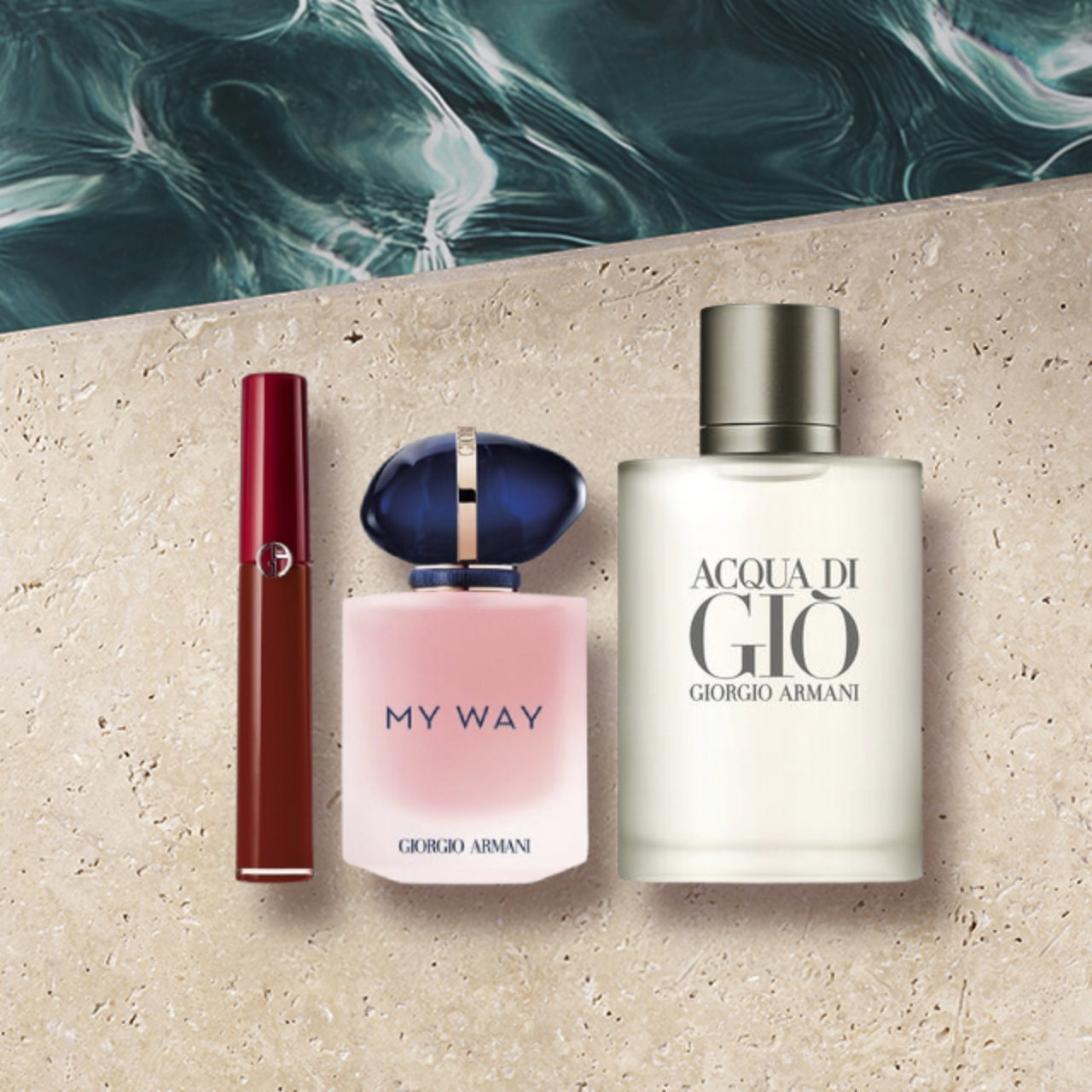 A flat lay of three Giorgio Armani beauty products: a red lip gloss, a "My Way" perfume bottle with pink liquid and an organic pink cap, and an "Acqua di Giò" perfume bottle with light liquid and a matte silver cap. They are arranged on a textured beige surface with blue water visible in the background