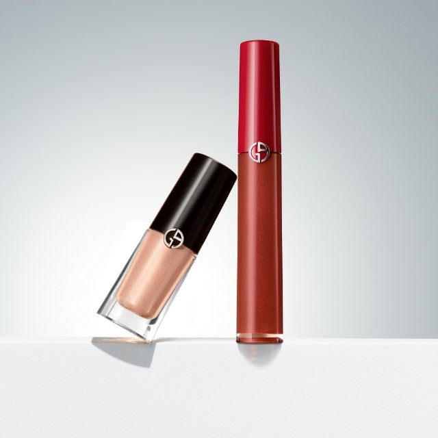 Two Giorgio Armani beauty products are displayed on a white surface against a light grey background: a clear rectangular bottle with a gold-toned liquid (likely a highlighter or eyeshadow) leaning against a taller, cylindrical red tube of lip gloss or liquid lipstick. Both feature the Giorgio Armani logo
