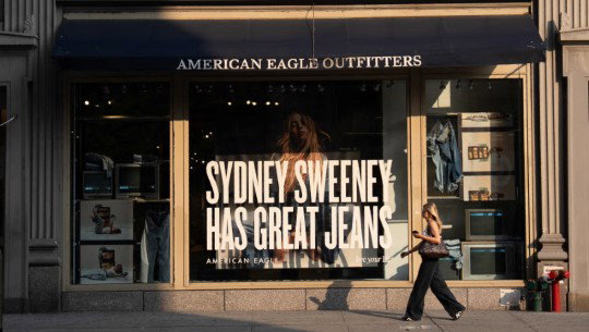 A woman passes an American Eagle story with a large poster in the window picturing Sydney Sweeney and the words "Sydney Sweeney Has Great Jeans"