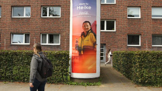 An outdoor ad for Coca-Cola says "Made by Heike" above the picture of a Coke employee holding a bottle of soda