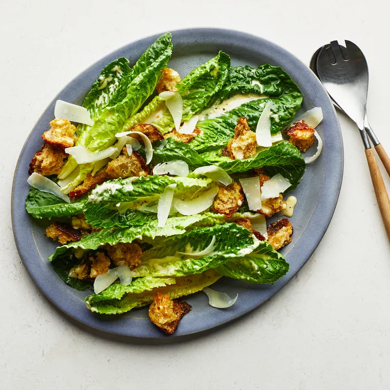 Gray platter of Caesar salad on a white backdrop with salad servers. 