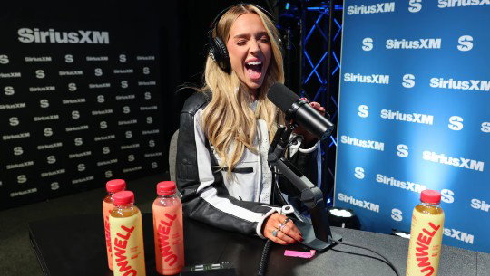 Alex Cooper at a SiriusXM microphone with bottles of Unwell branded drinks on the table