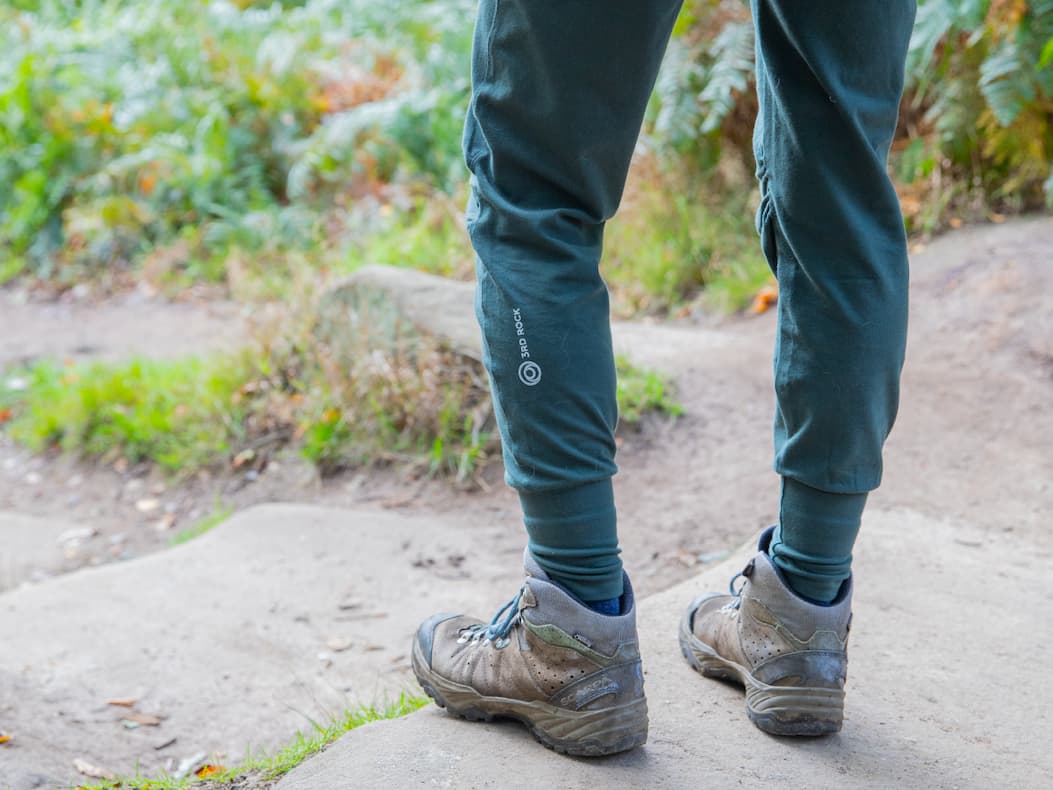 Accessible description: A close up of a climber's leg as they ascend a boulder. They are wearing the new Bataboom trousers in the dark green scarab colour. The photo frames a 3RD ROCK logo on the left calf