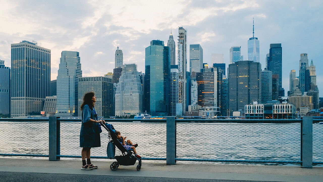 A mother pushes her baby in a pushchair along the waterfront with the New York City skyline behind