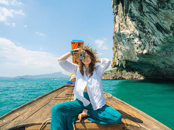 Frau mit Kamera vor dem Gesicht sitzt auf einem thailändischen Boot.