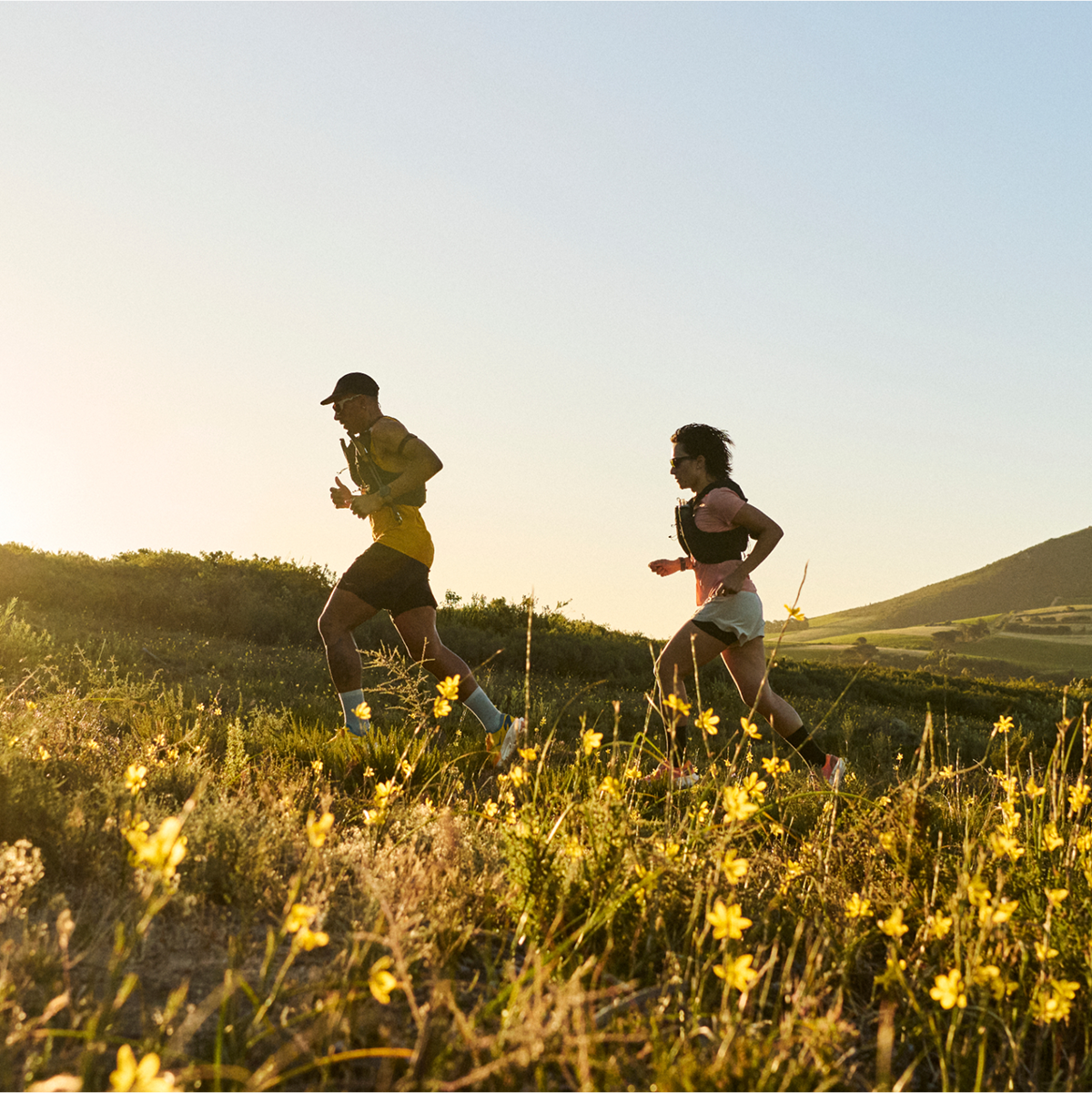An image of two people running through a field of grass and flowers with the sun setting and a mountain in the background.