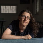 A woman with long, dark hair and glasses leans with one arm against the arm of a couch.