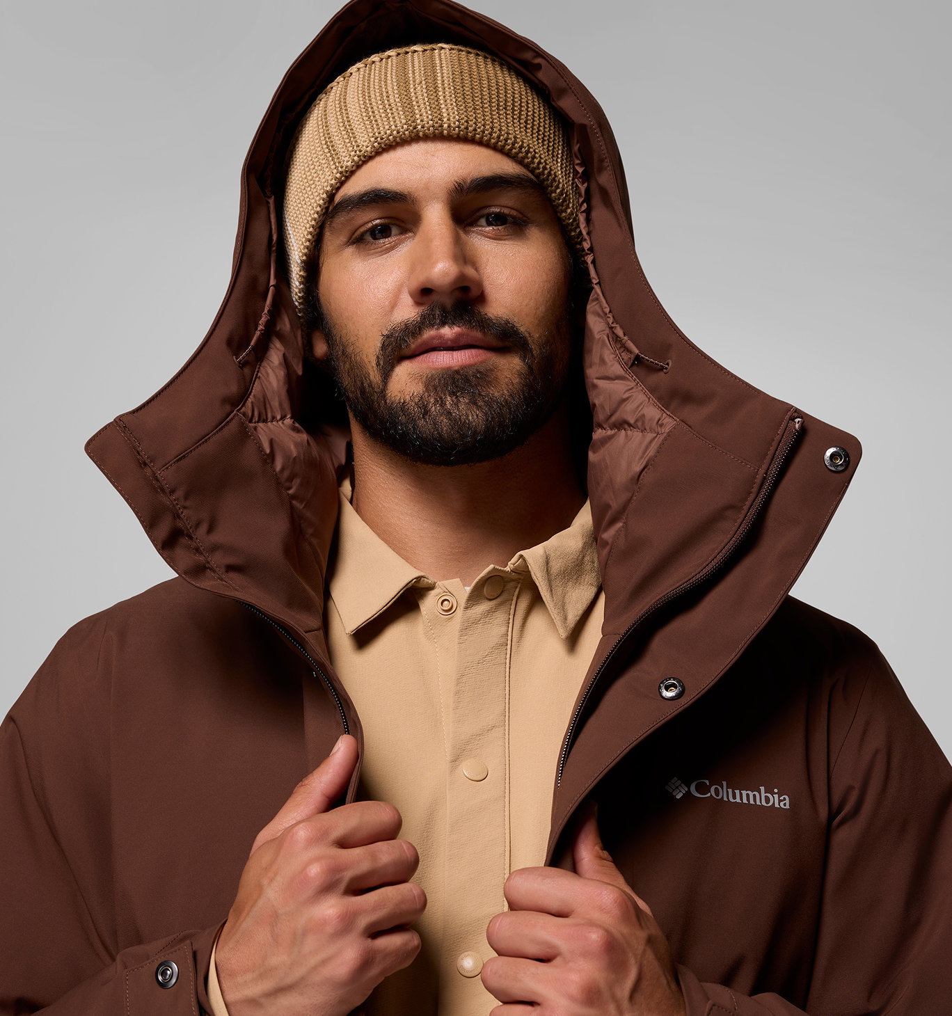 A man wearing a beanie, tan shirt, and brown jacket with the hood up.