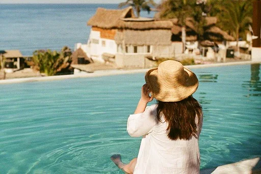 A woman with long brown hair and a straw hat dips her feet in an infinity pool as she looks out at the sea