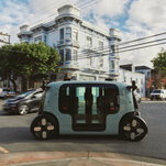 A boxy green vehicles passes by a white Victorian-style building.