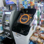 A Bitcoin A.T.M., with a black and orange screen, next to a gray, traditional A.T.M. Both stand in the midst of shelves of packaged pastries and other items.