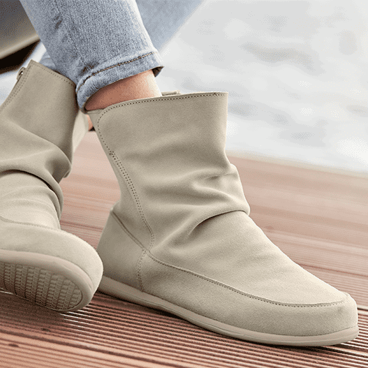 Eine Person trägt beige, knöchelhohe Stiefeletten und helle Jeans auf einem Holzboden.