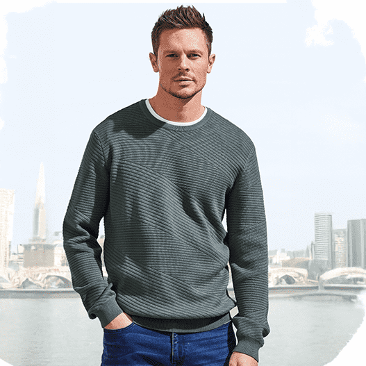 Ein Model trägt einen dunkelgrünen Strickpullover und blaue Jeans. Im Hintergrund eine Stadt mit Fluss und Himmel.