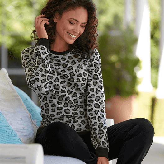 Ein Model mit gelockten Haaren sitzt auf einem Stuhl und trägt ein graues Leopardenmuster-Oberteil und schwarze Hose, mit Kissen im Hintergrund.