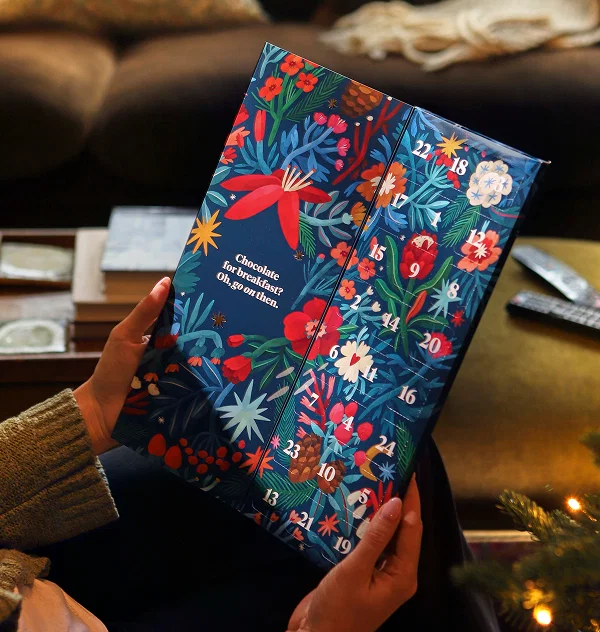 Someone sat in a cosy living room holding a Bloom & Wild advent calender in a fun festive print, with numbered doors on one side and text on the other that reads 