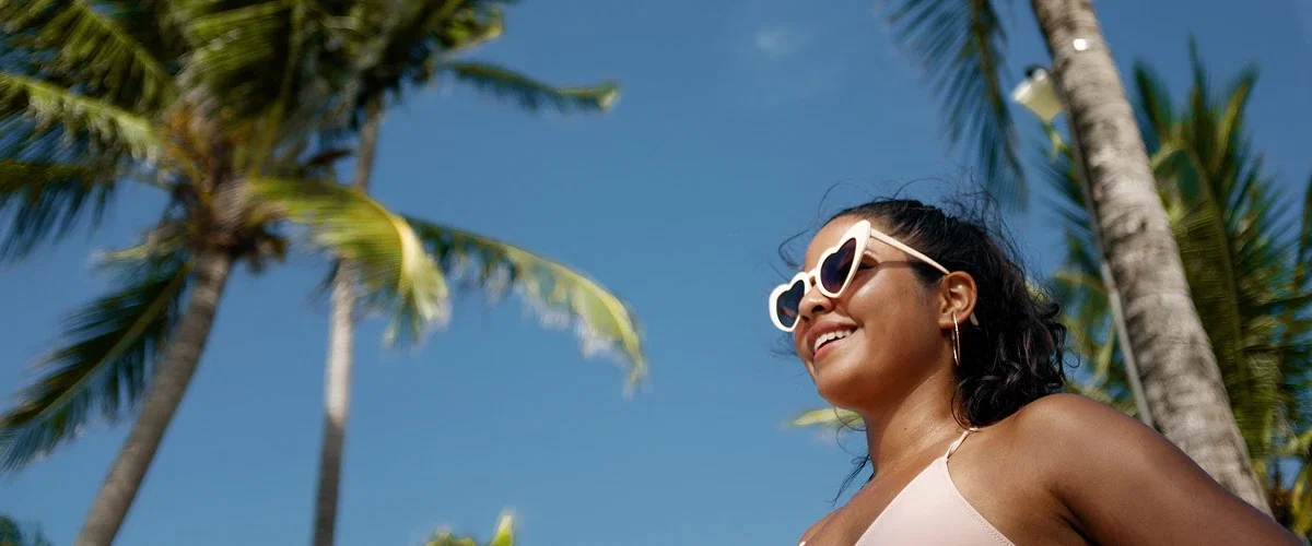 A woman in a pink bathing suit and sunglasses smiles with palm trees in the background.