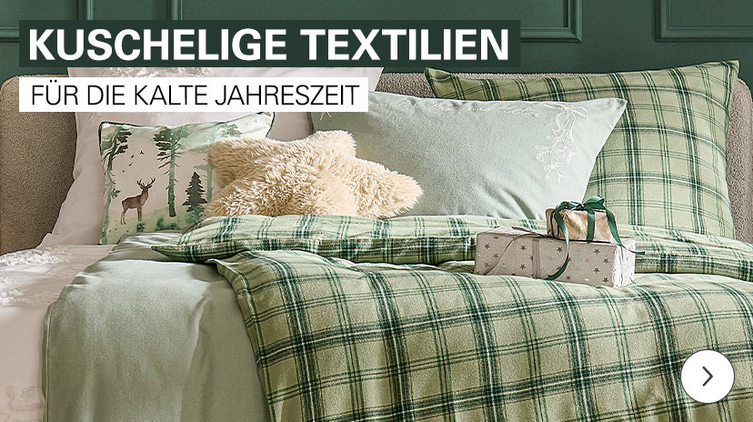 Ein Bett, bedeckt mit hellgrüner und grün-karierter Bettwäsche sowie weichen Zierkissen, darunter eines mit Hirsch-Motiv und ein flauschiges. Auf der Decke liegen zwei verpackte Geschenke. Oben steht der Text 'KUSCHELIGE TEXTILIEN FÜR DIE KALTE JAHRESZEIT'.