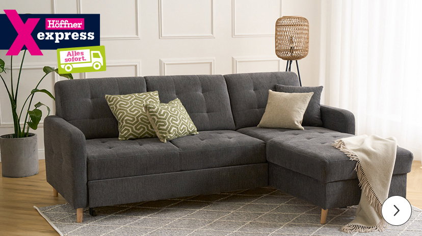 Dunkelgraues Ecksofa mit Ottomane und Holzkufen, auf dem helle und olivgrün gemusterte Kissen sowie eine beigefarbene Decke liegen. Oben links das 'Höffner Express' Logo mit dem Hinweis 'Alles sofort'.