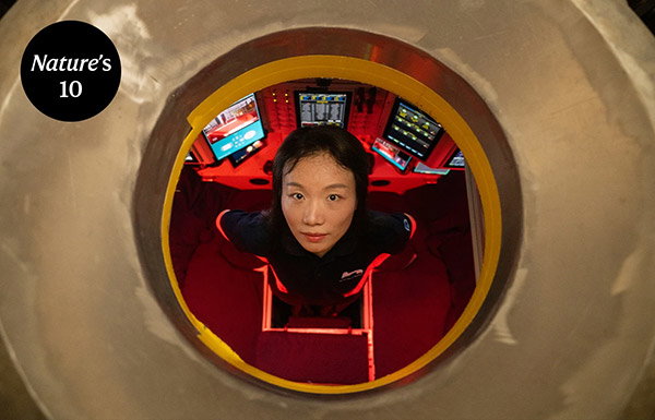 Portrait of Mengran Du standing in a red-lit space with a bank of computer screens, looking up through small circular hole in a submarine.