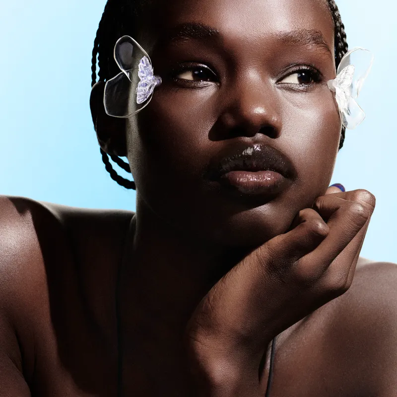 A close-up shot of a dark-skinned model with glowing skin wearing makeup accessories on the outside of both eyes