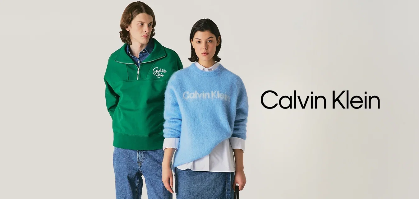 Mężczyzna w zielonym swetrze z polo i dżinsach stoi obok kobiety w niebieskim swetrze Calvin Klein, w dżinsowej spódnicy, trzymającej czarną torebkę.
