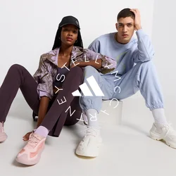 Dwie modelki siedzą na jasnej powierzchni. Jedna ma na sobie wzorzystą brązową kurtkę, fioletowe legginsy i różowe sneakersy. Druga ubrana jest w jasnoniebieski dres i białe sneakersy.