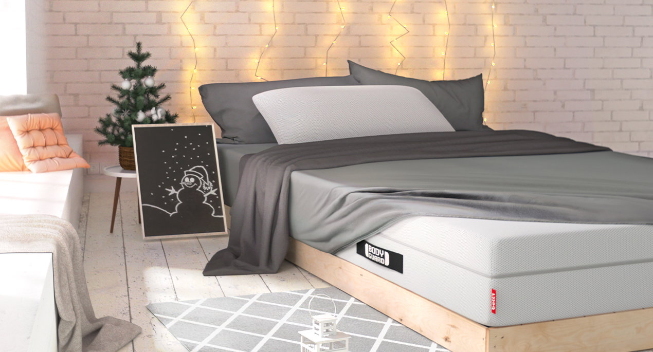 In einem weihnachtlich dekoriertem Raum steht ein Bett mit BODYGUARD Matratze, darauf liegt das BODYGUARD Visco-Kissen. An der Wand hängen Lichterketten.