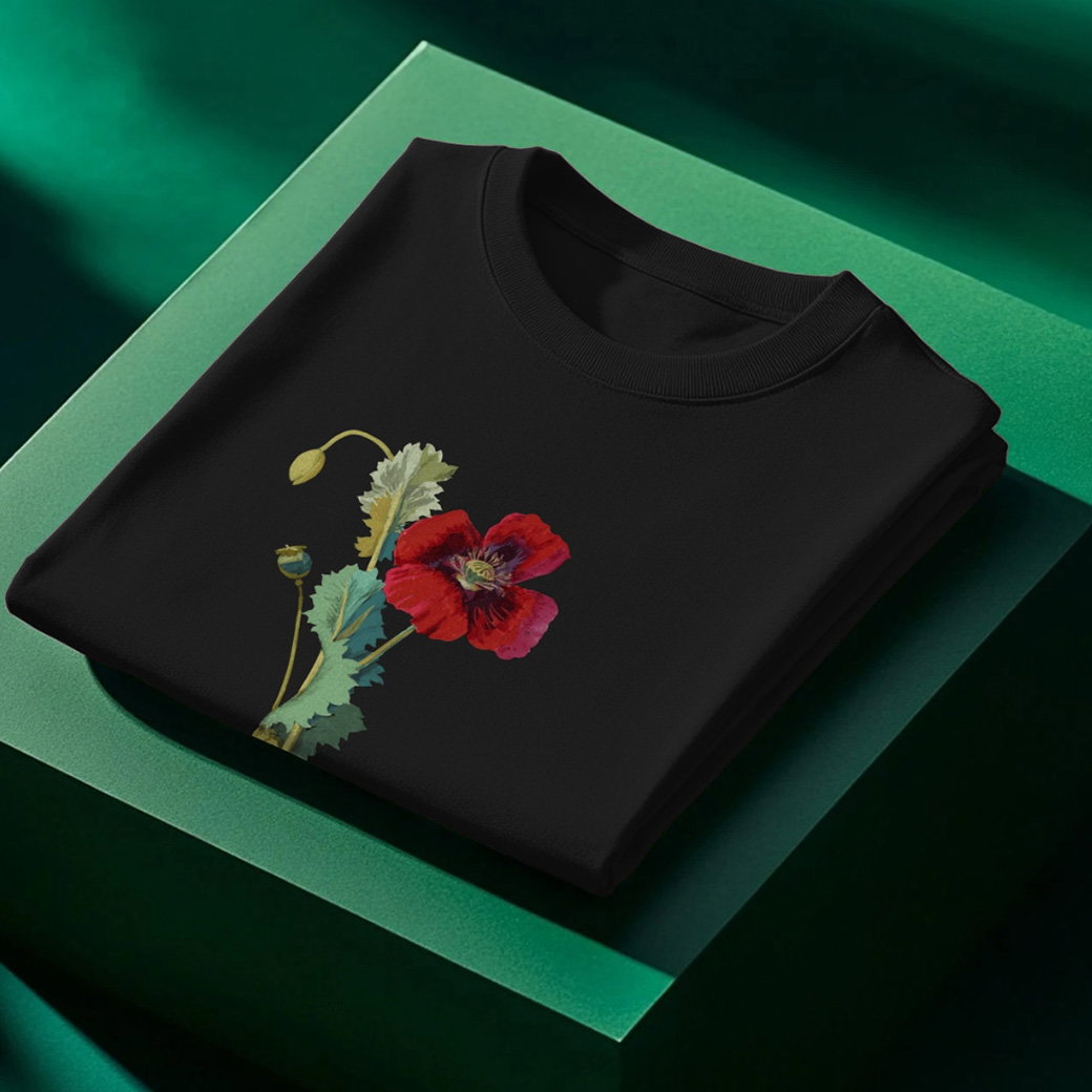 A folded unisex cotton t-shirt in black featuring Mary Delany’s Opium Poppy design on a green block. – Shop Now. 