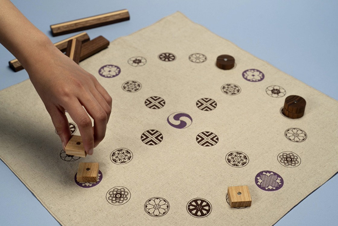 A contemporary reinterpretation of a traditional Korean yutnori board, featuring traditional motifs, with playing pieces on the board and wooden sticks behind, as a hand moves a piece.