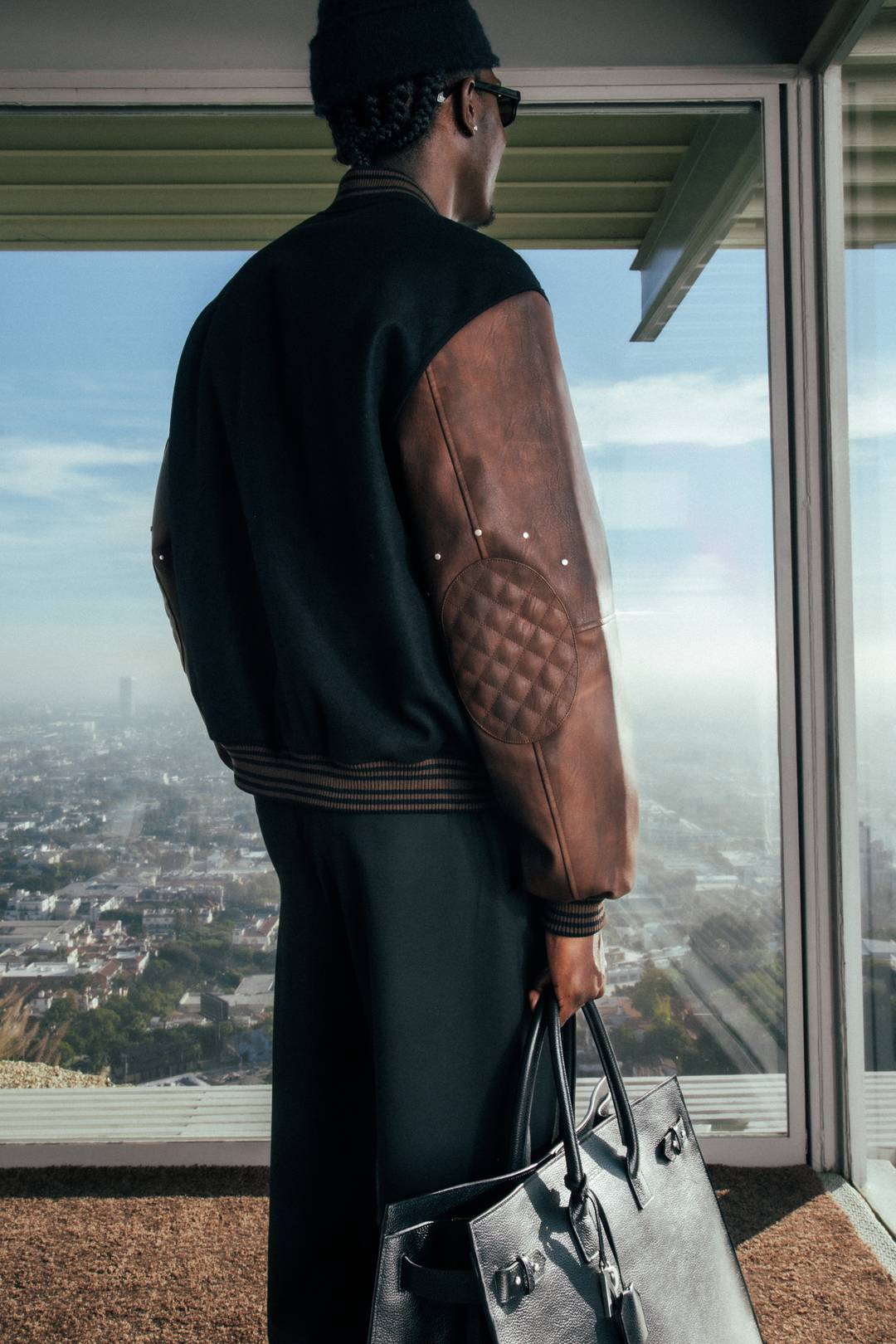 Model wearing the Varsity Bomber in Jet Black and holding the HTN Leather Bag in Black